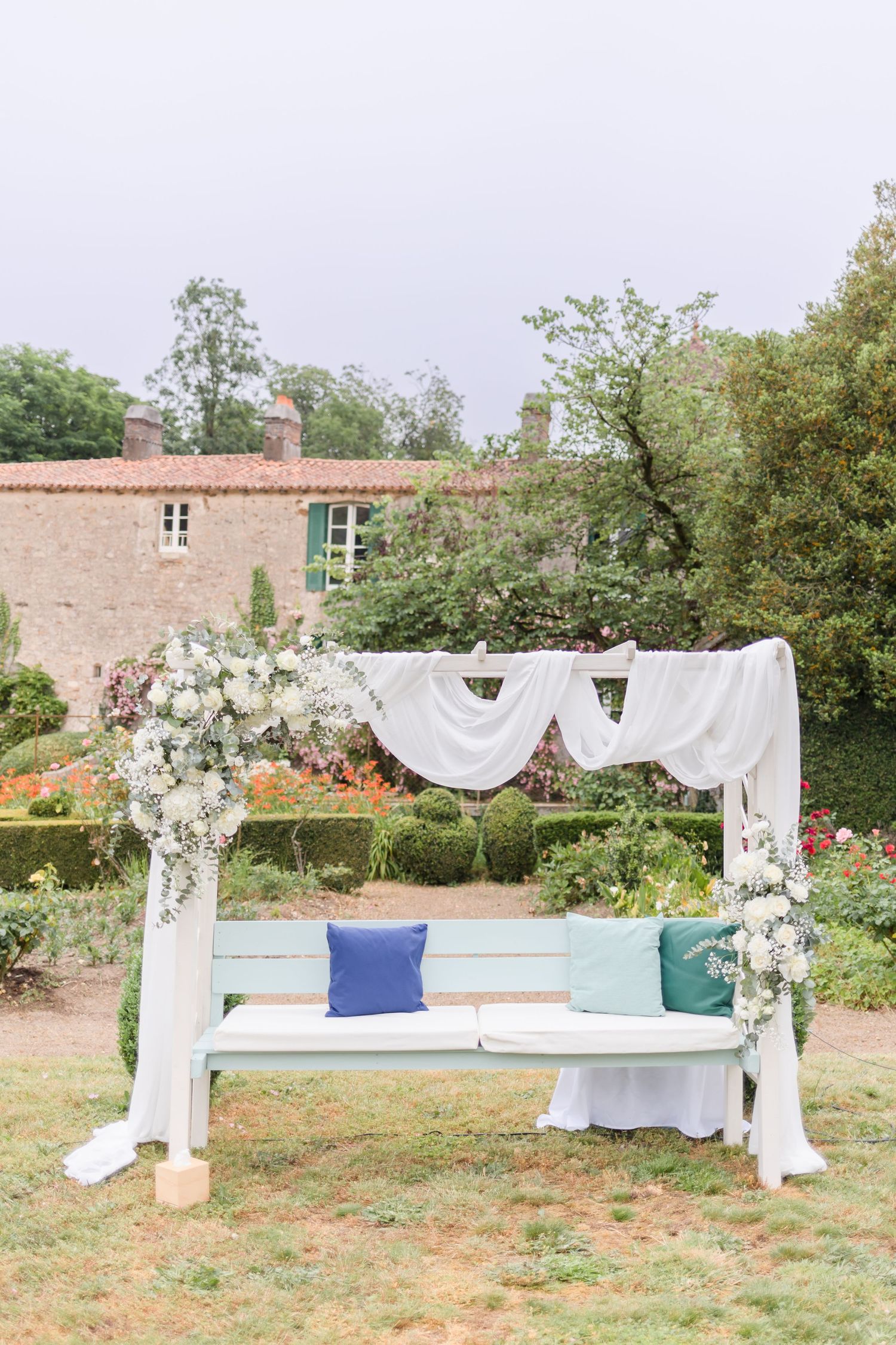Elegant white draped wedding arch with floral arrangements and blue cushioned bench in a garden setting.