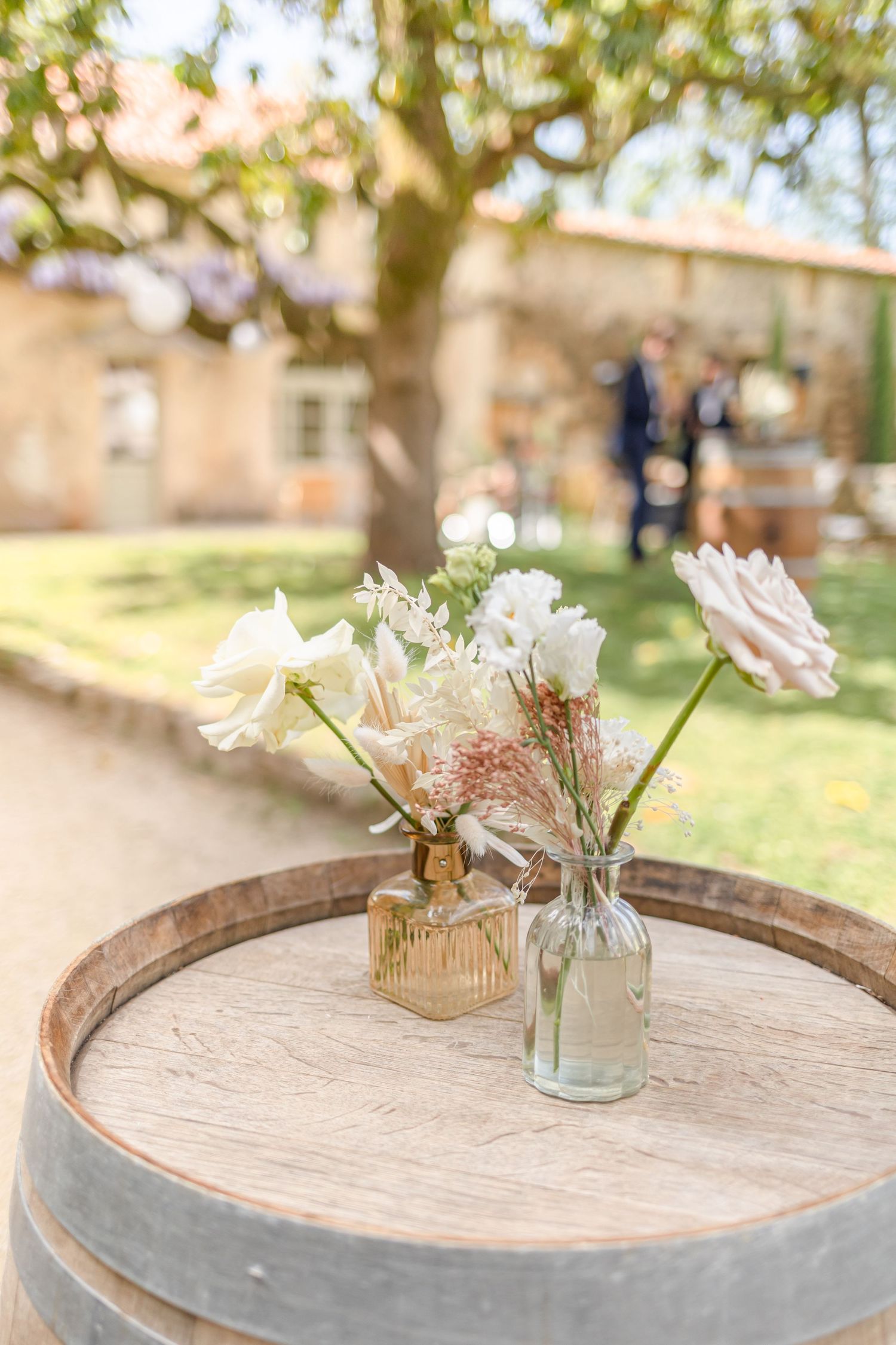 Rustic wooden barrel table displays delicate floral arrangements in glass vases with soft natural lighting.