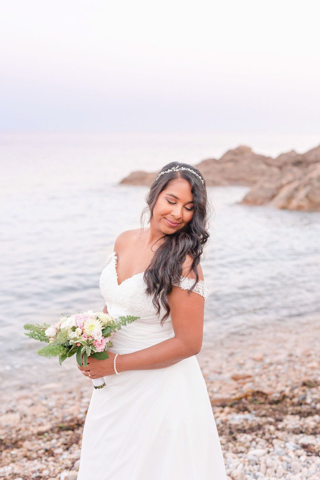 A bride in an off-shoulder gown holds her bouquet while posing on a rocky beach.