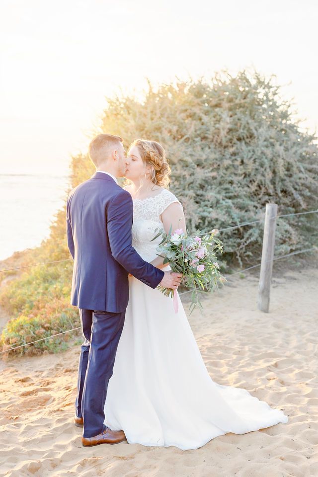 A newlywed couple embracing on a sandy beach with greenery and ocean waves in the background.
