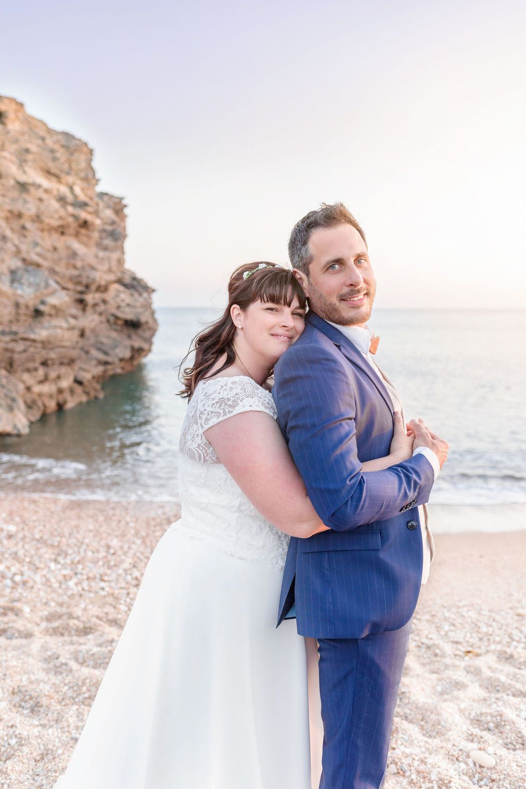A couple in wedding attire poses together on a beach with coastal rocks in the background.