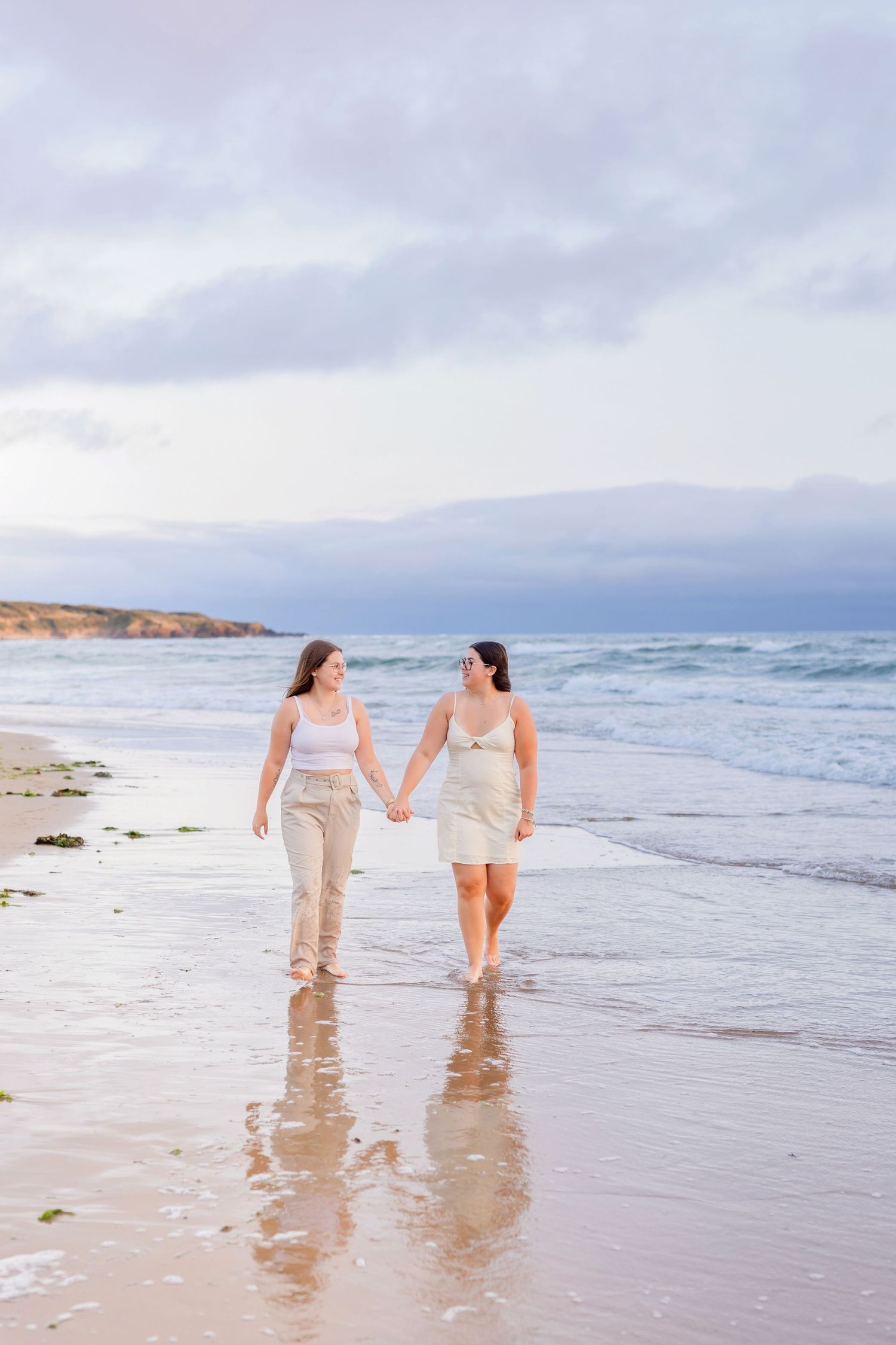 Two people walk hand in hand along the shoreline at sunset with waves lapping at their feet.