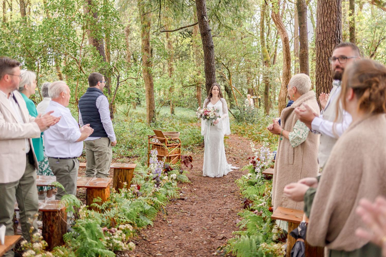 Rustic woodland wedding ceremony with wooden benches and ferns lining a dirt path.