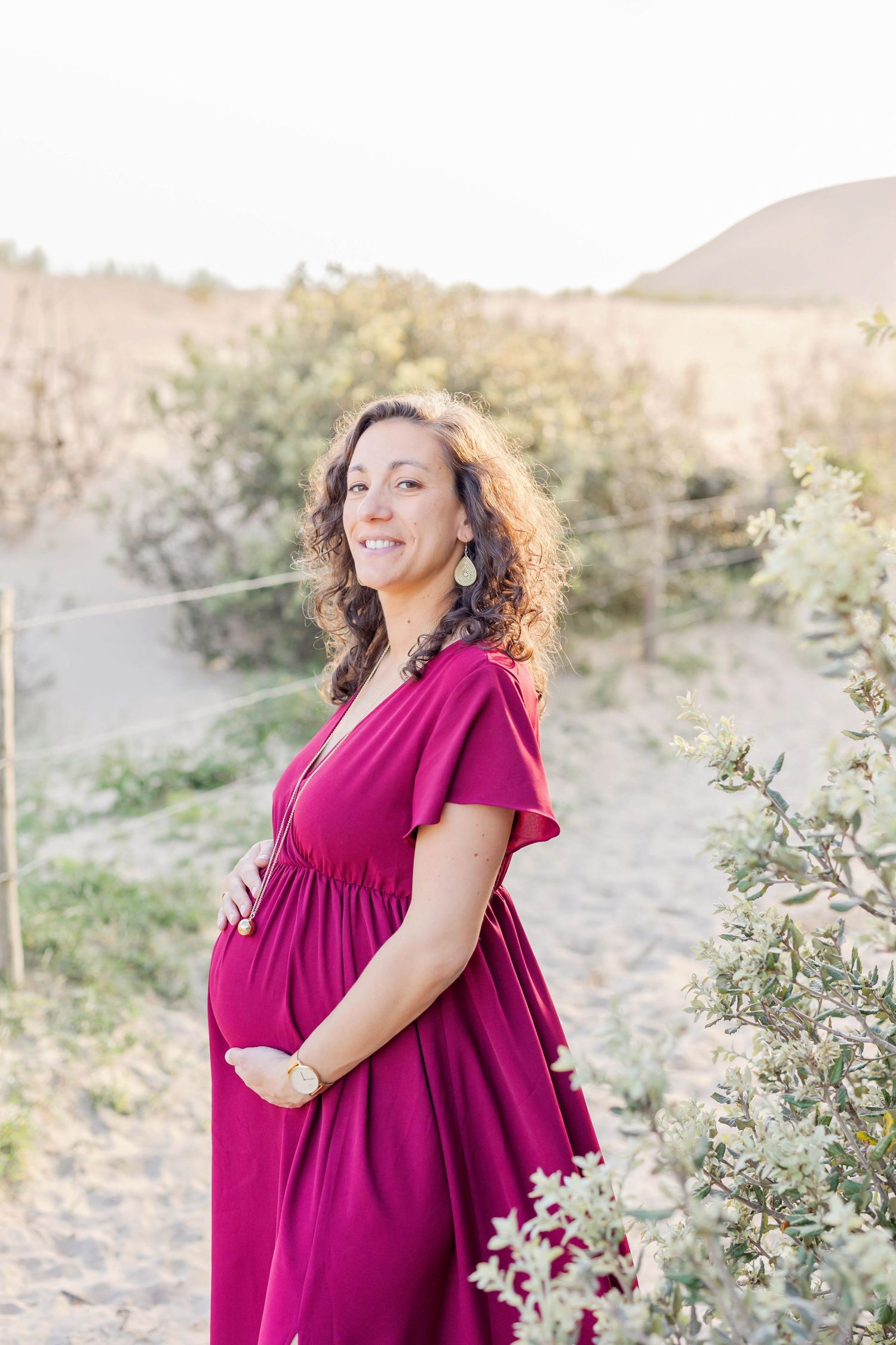 Expectant mother in burgundy dress poses against desert landscape during golden hour photoshoot.
