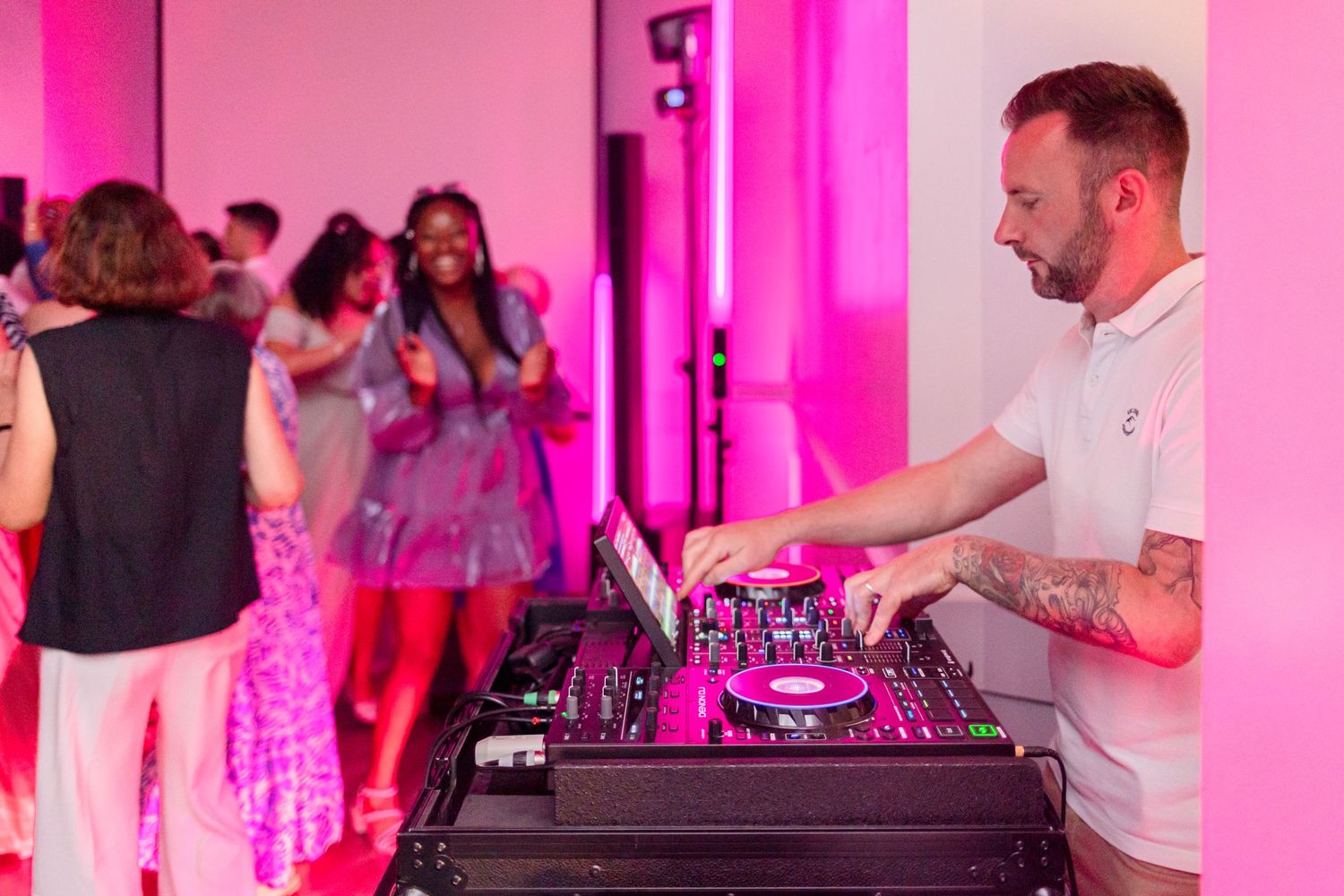 DJ performs at wedding reception with pink lighting while guests dance in background.