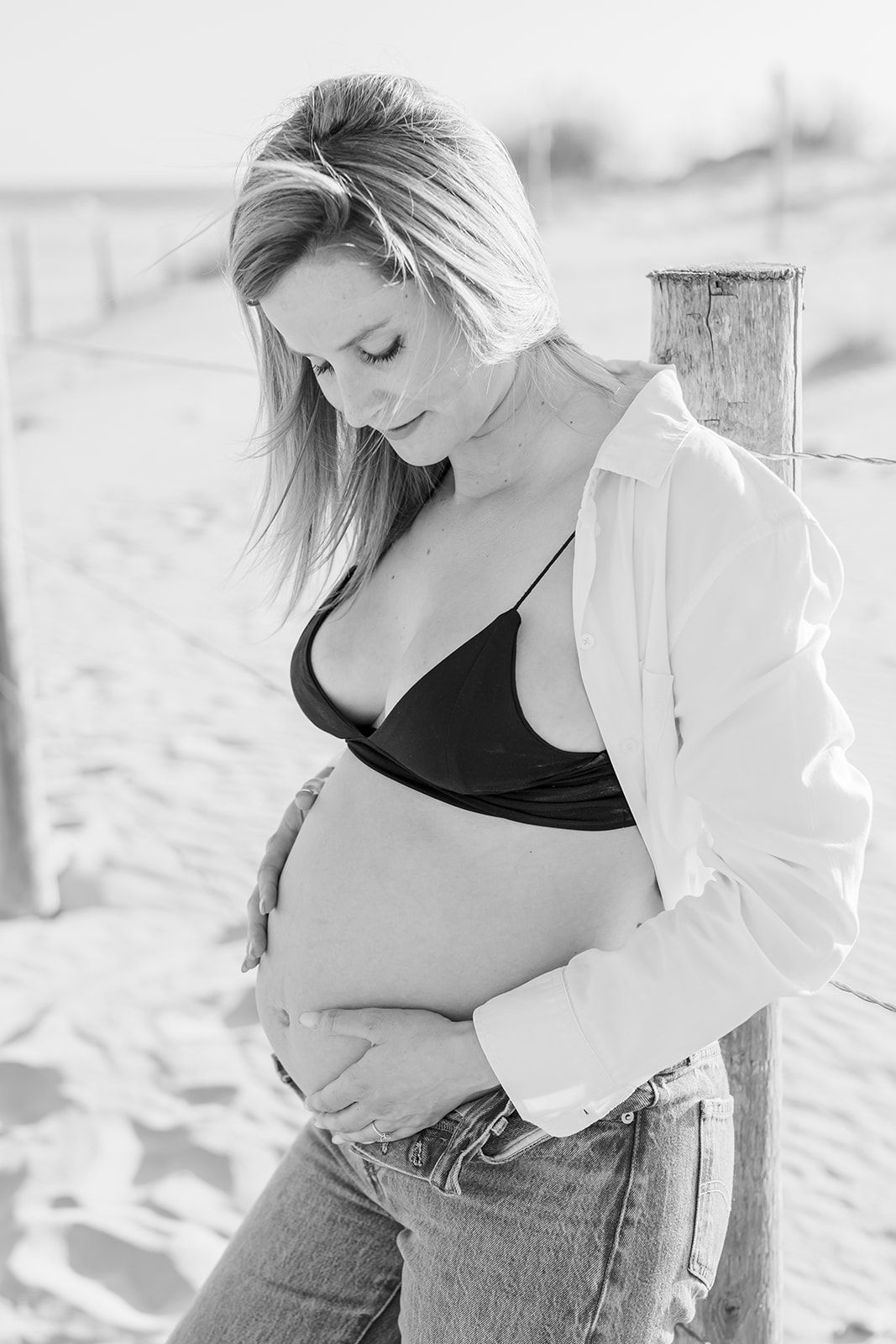 Black and white maternity photo series showing a pregnant woman in casual attire posing outdoors on a beach.