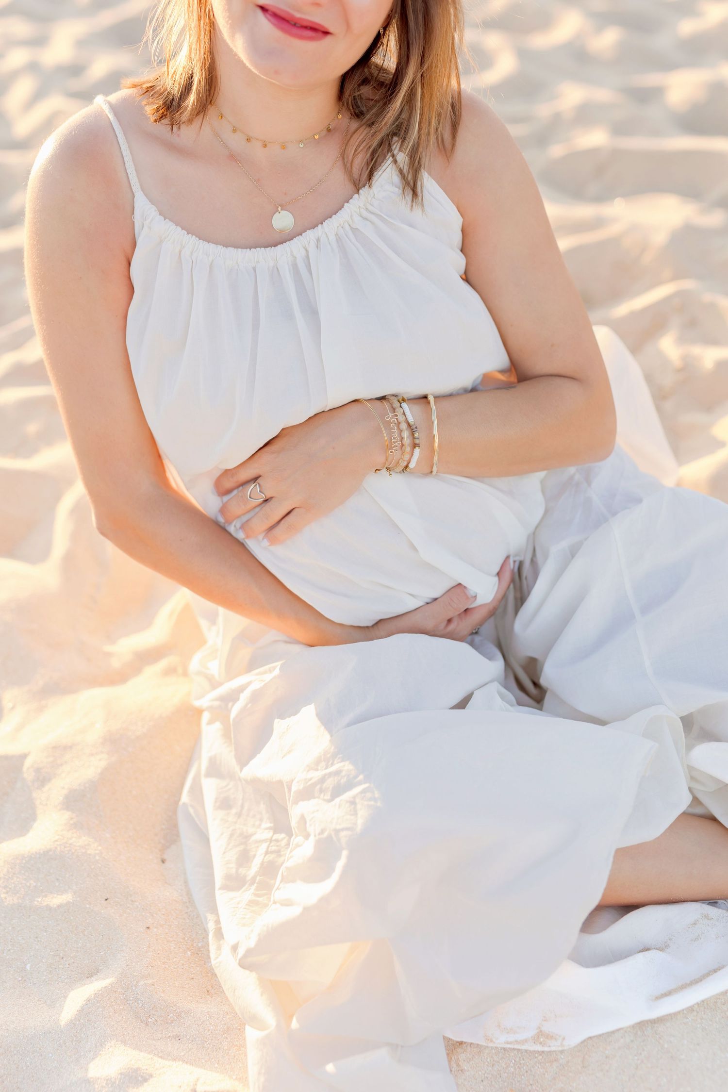 A person in white clothing sits on sandy beach wearing jewelry and bracelets during golden hour.