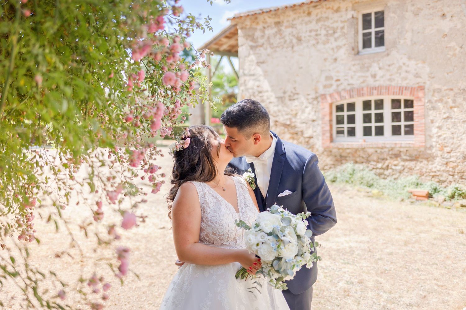 Bride and groom pose for romantic photos near blooming pink flowers against stone villa backdrop.