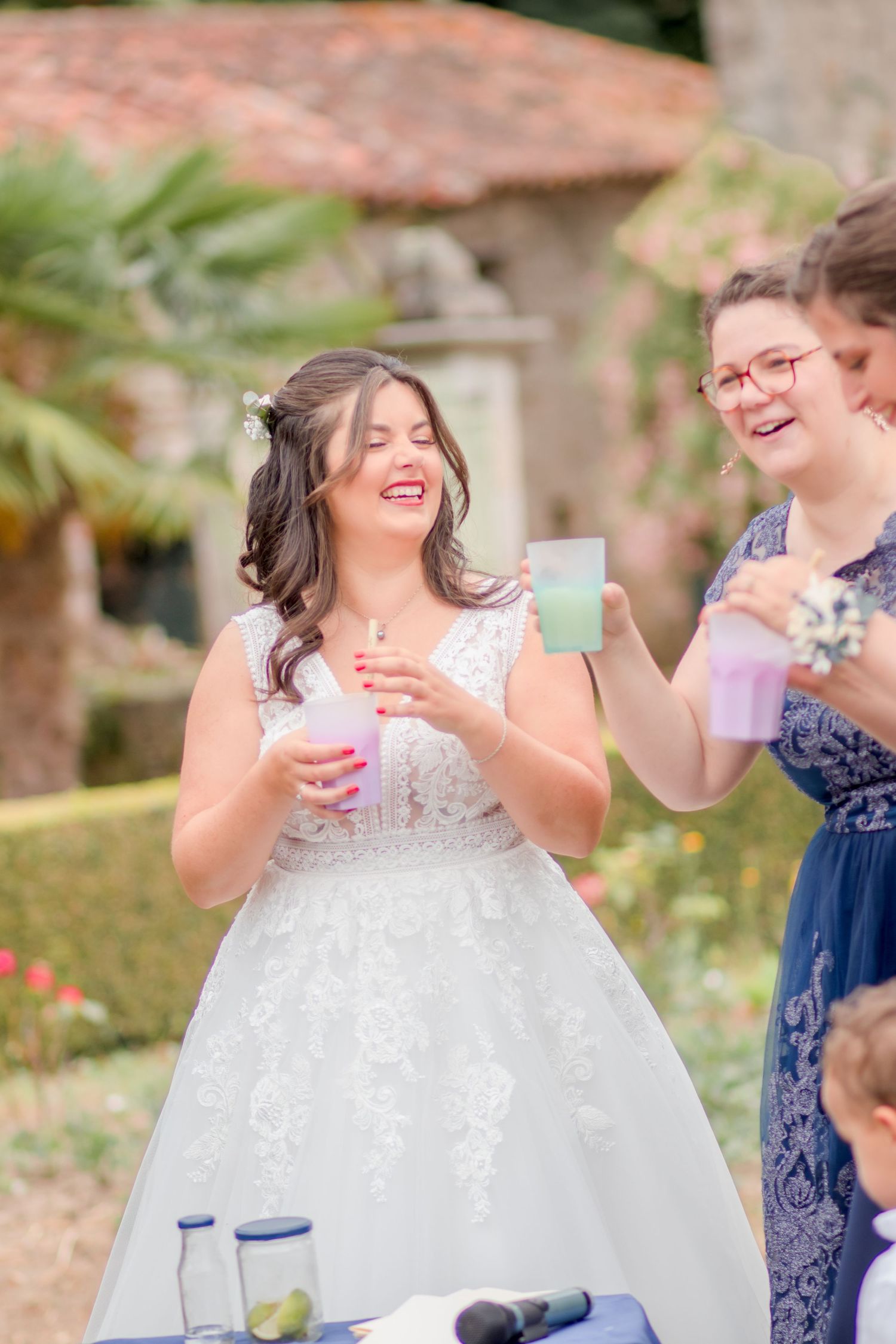 A bride in a white dress shares drinks and laughter during outdoor wedding celebrations.