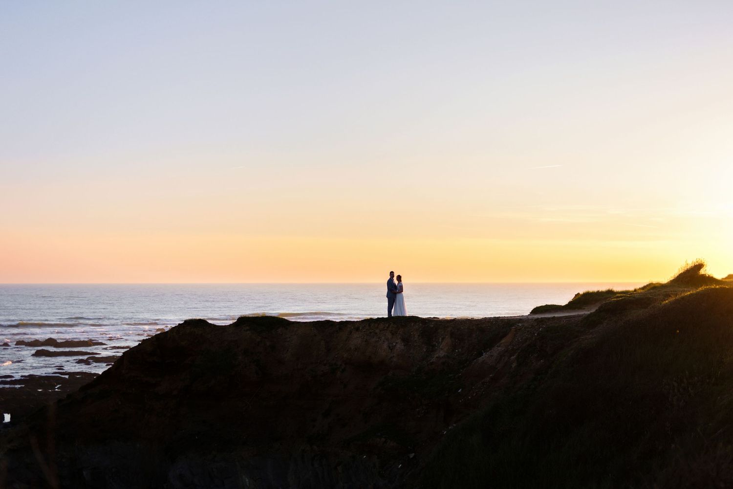 Silhouettes of a couple standing on coastal cliffs at sunset.