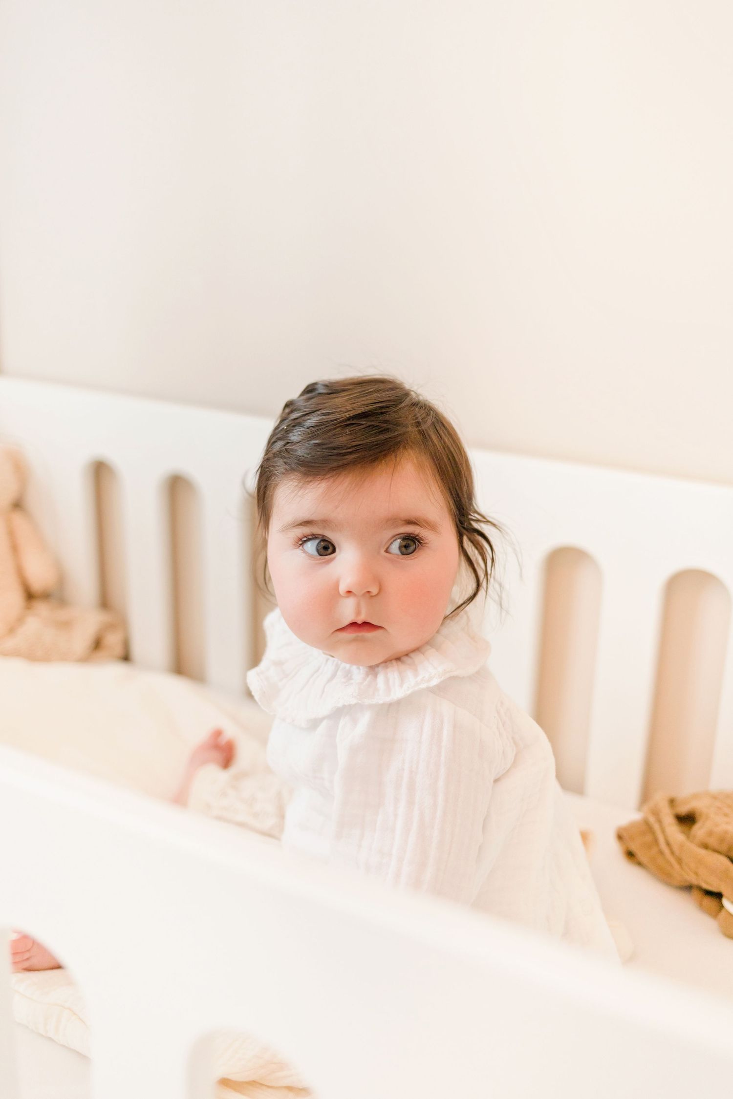 Baby rests against white crib railing in soft white clothing.