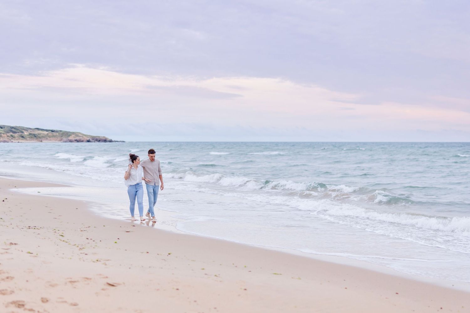 Couple walking along shoreline at sunset with ocean waves in background.