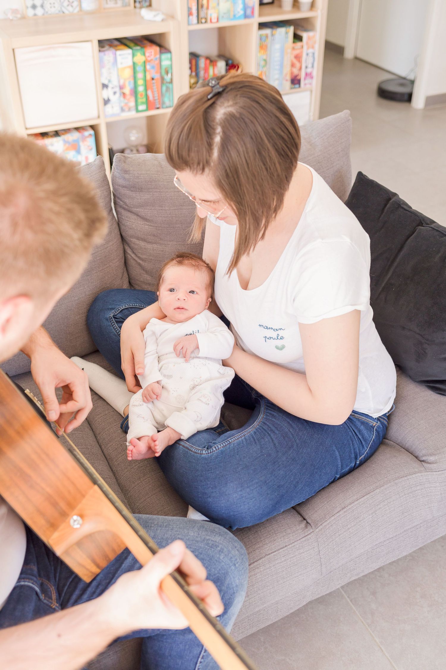 A baby on a grey couch watching someone play a wooden ukulele in a bright living room.