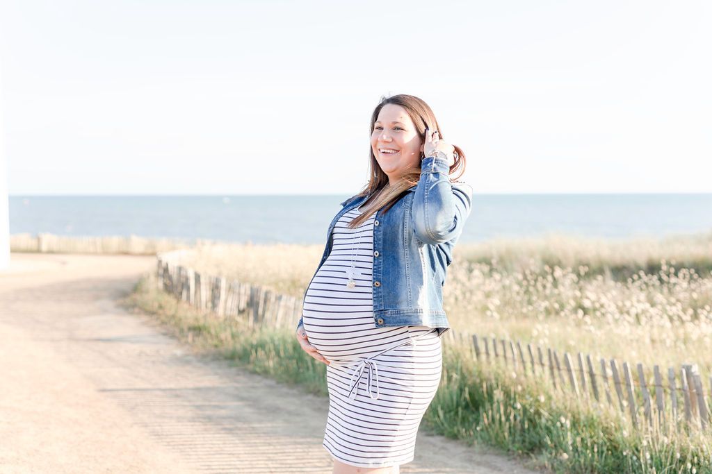Expectant figure in a striped dress and denim jacket poses by the ocean.