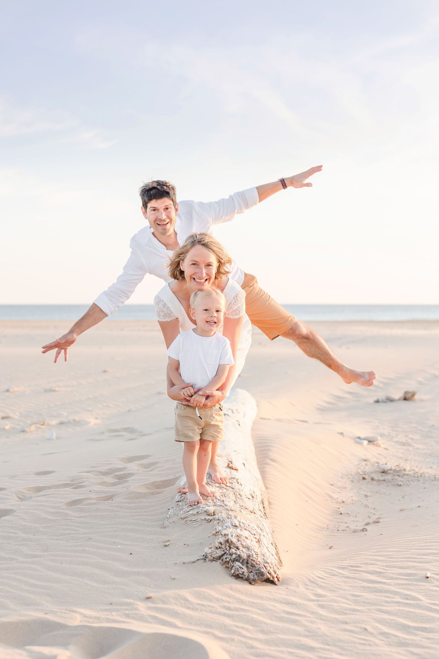 Three people playfully balancing with outstretched arms on a sandy beach ridge at sunset.
