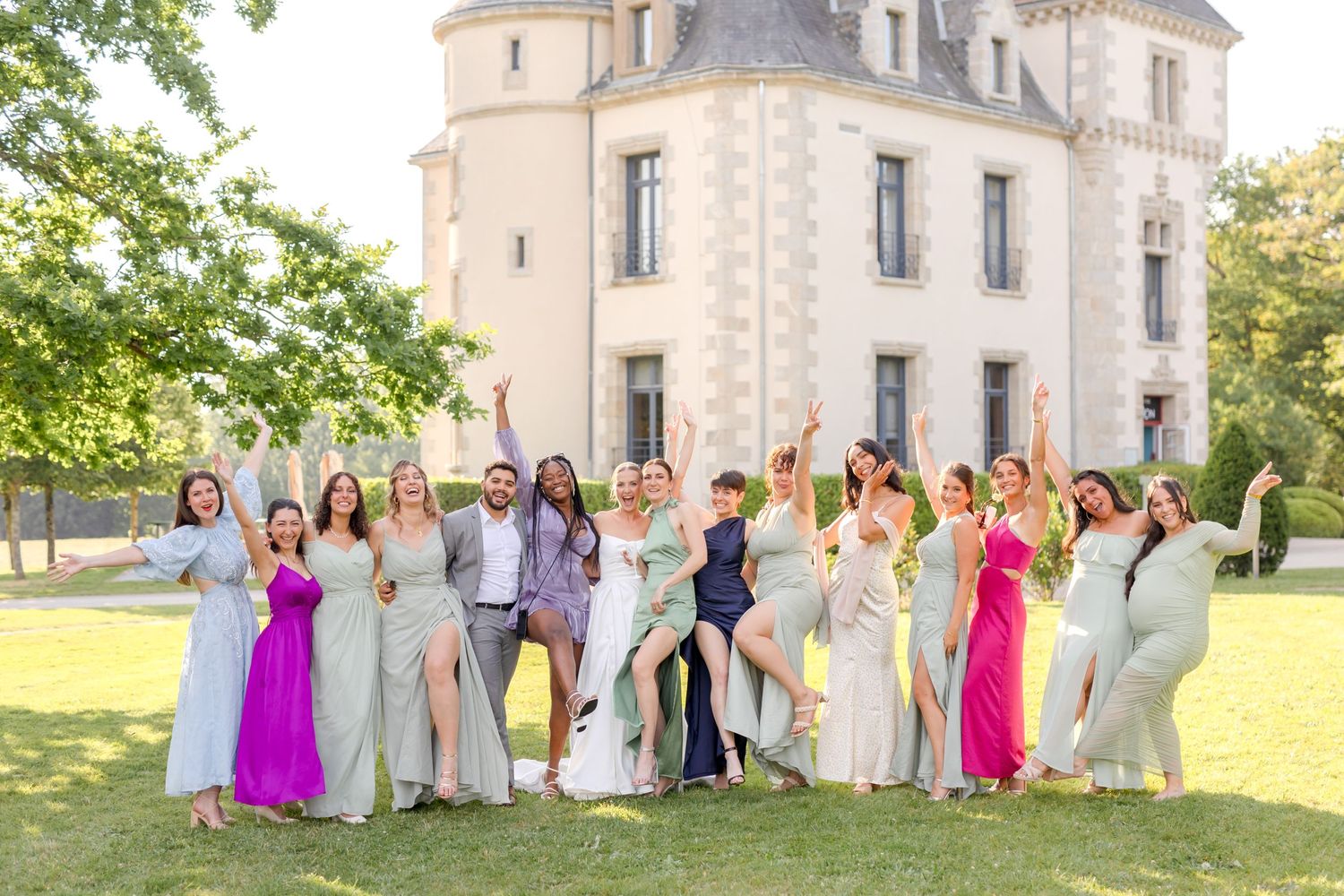 Large wedding party in vibrant dresses poses for group photo in front of elegant castle.