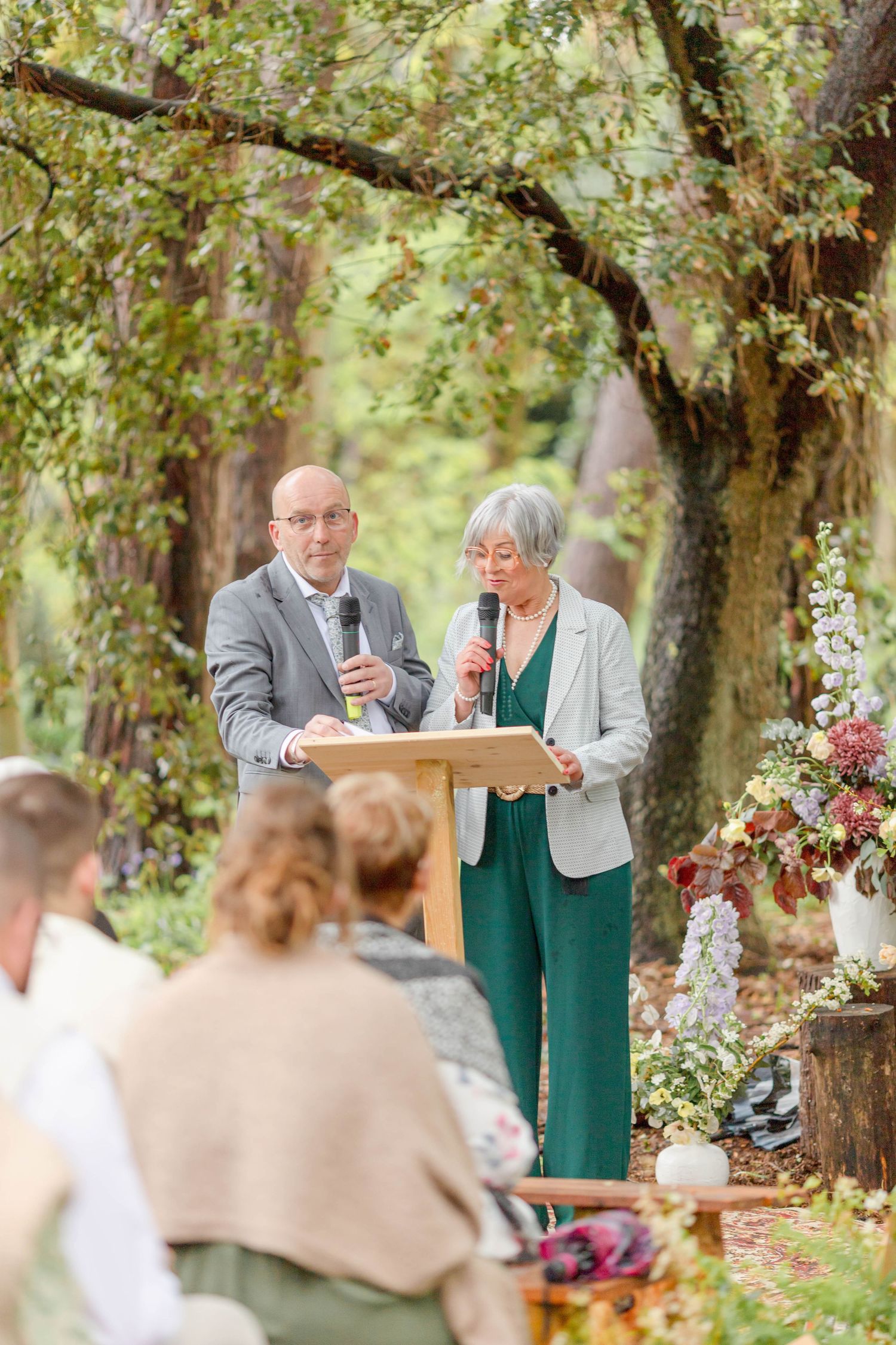 Outdoor woodland wedding ceremony with floral decorations and guests seated on wooden benches.