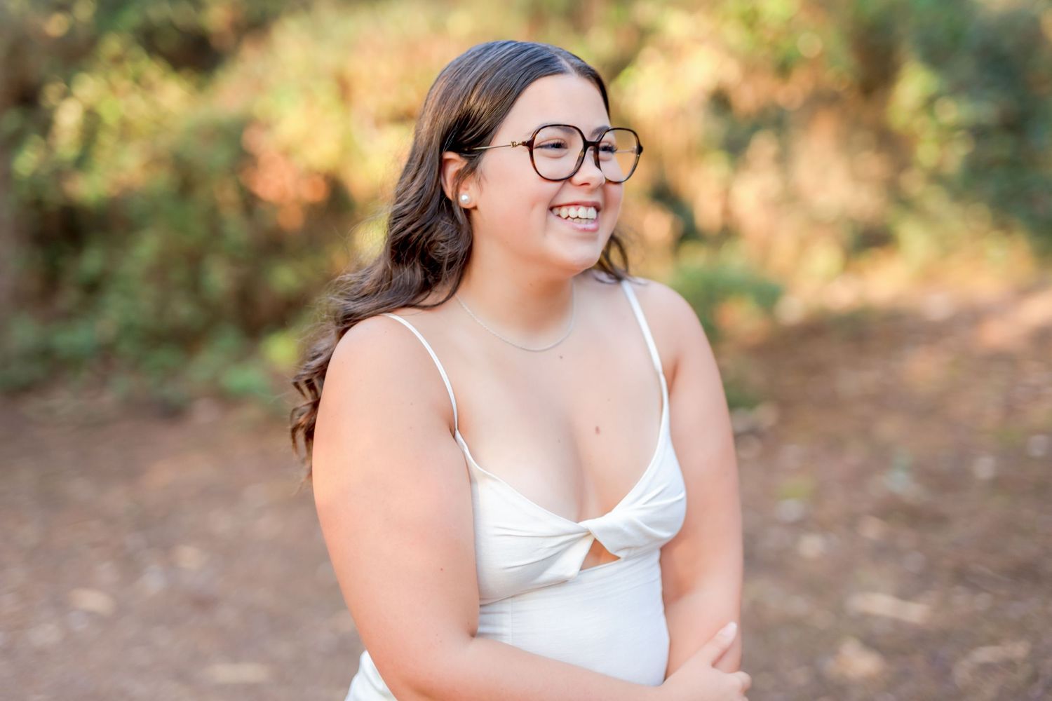 A smiling person wearing glasses and a white dress poses outdoors in a natural setting.