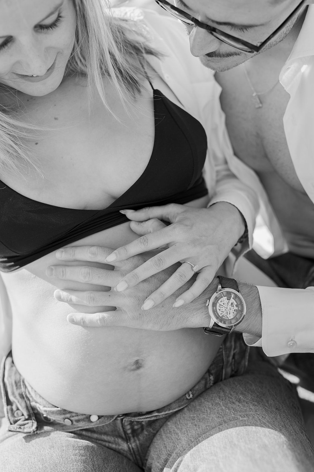 Black and white close-up photograph of hands tenderly resting on a pregnant belly, with a watch visible on one wrist.