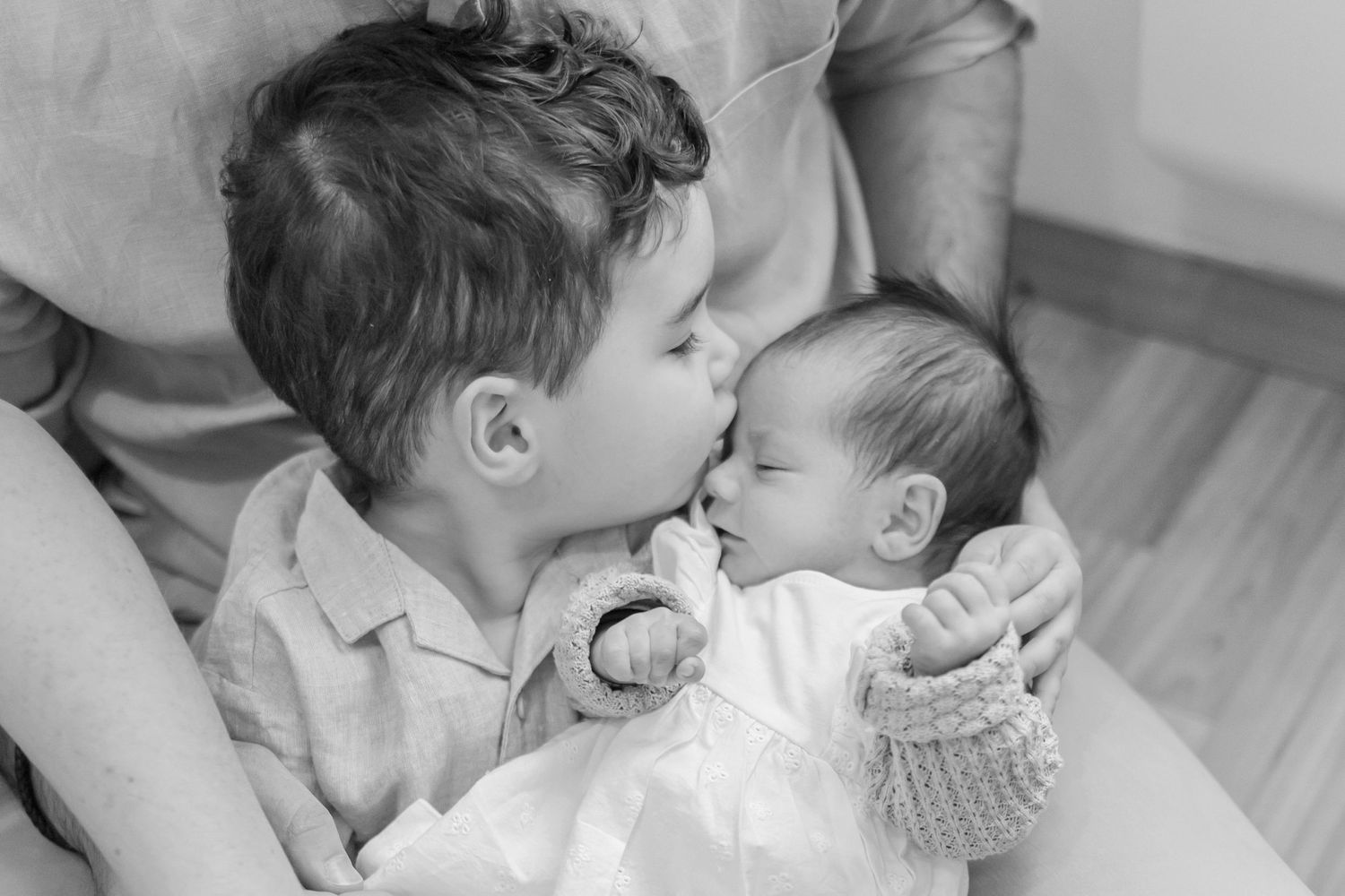 A tender black and white photo showing a toddler gently kissing a newborn baby in white clothing.