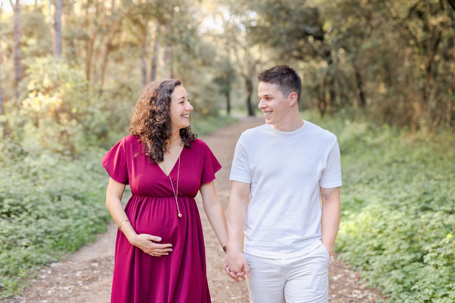 A couple embraces on a sunlit nature path wearing burgundy and white outfits during a maternity photoshoot.