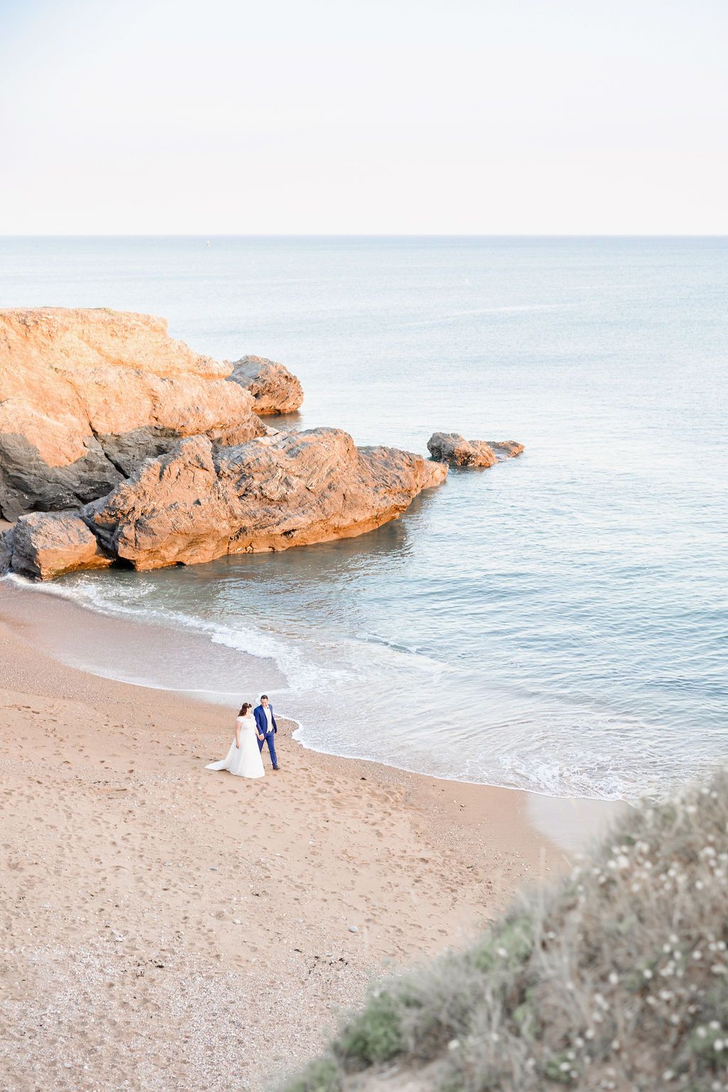 A romantic aerial view of a wedding couple on a pristine beach with dramatic coastal rocks.
