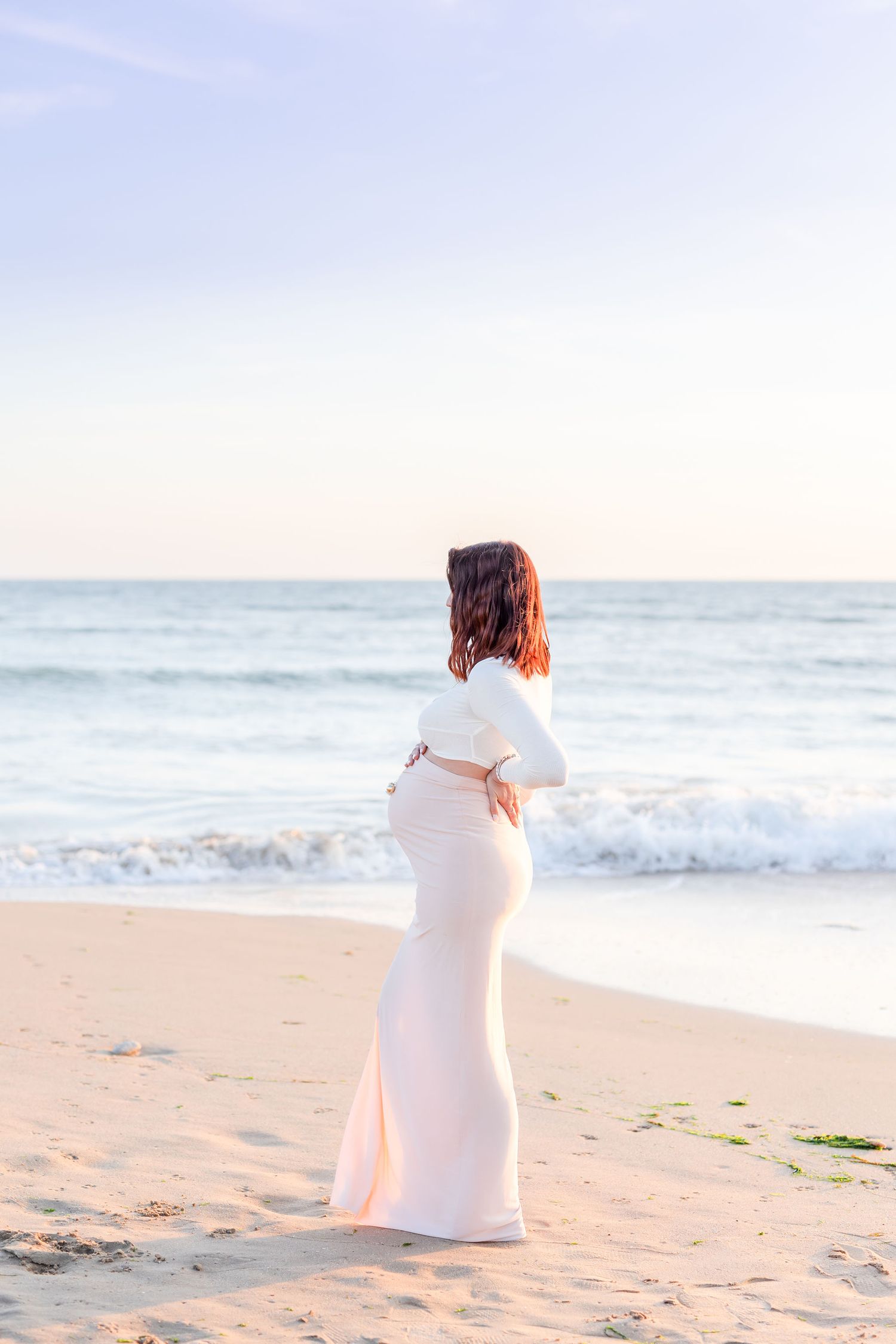 A figure in flowing white dress walks along the shoreline at golden hour.