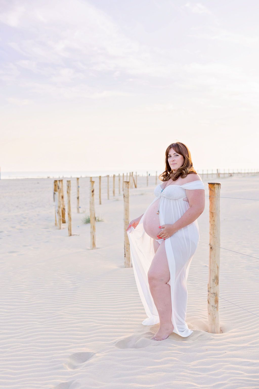 Dreamy maternity beach photo with wooden posts and white flowing dress at dusk.