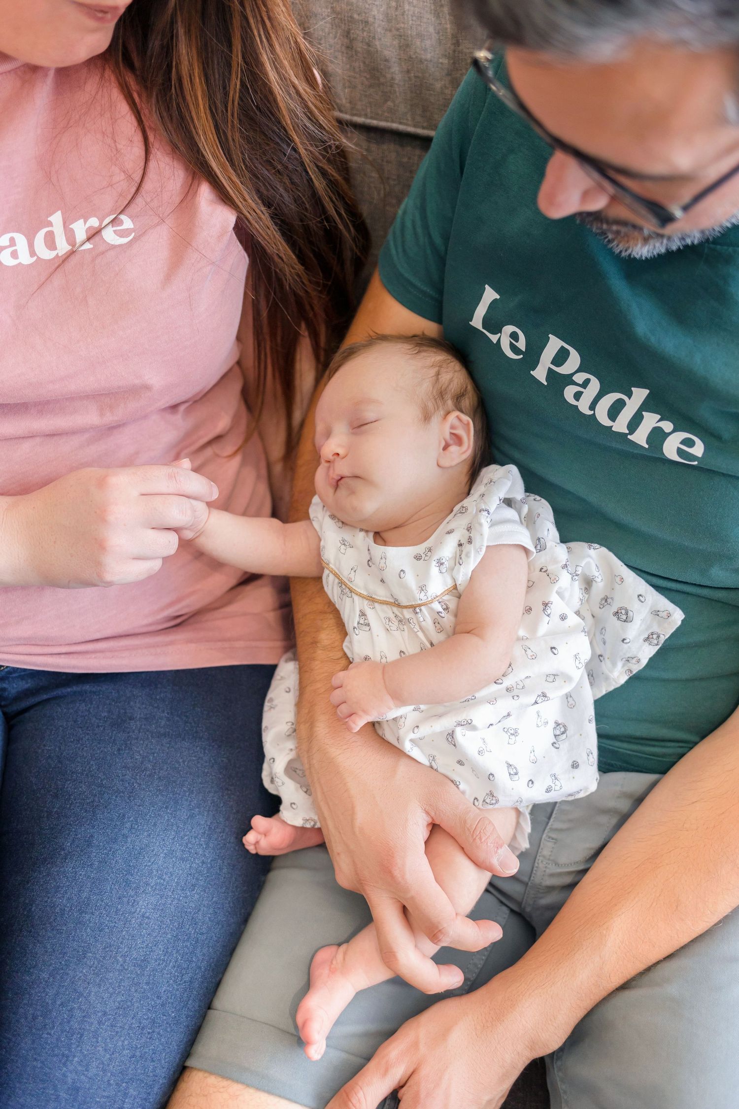 Tender moment with sleeping infant in white dress being held on couch.