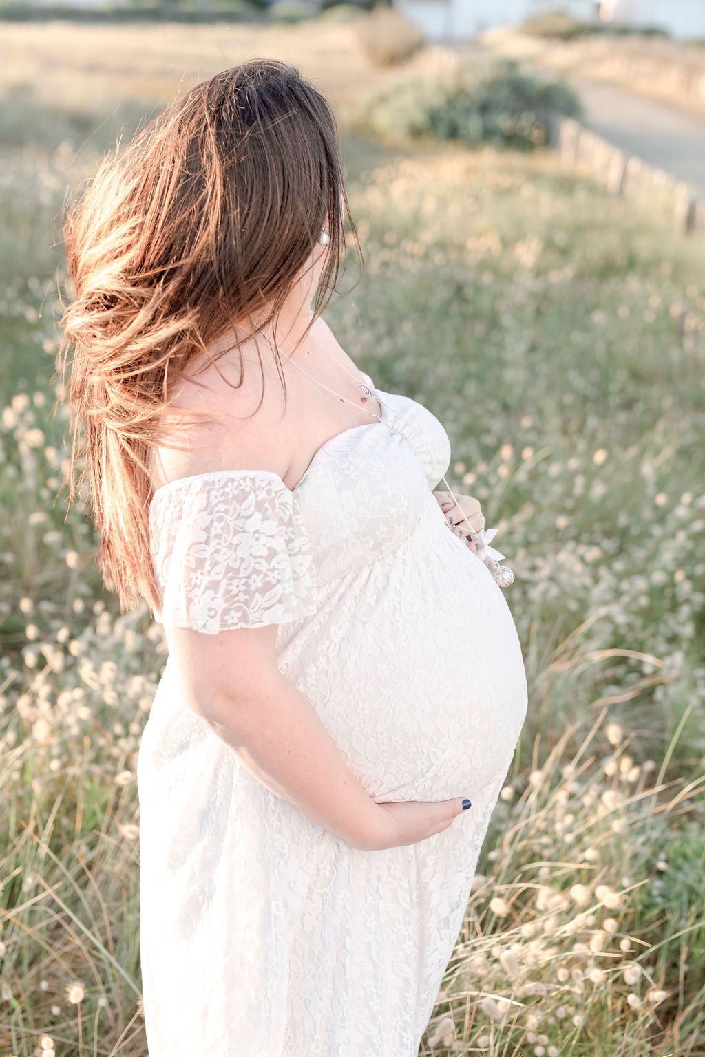 Side view maternity photo capturing flowing white dress against coastal meadow.