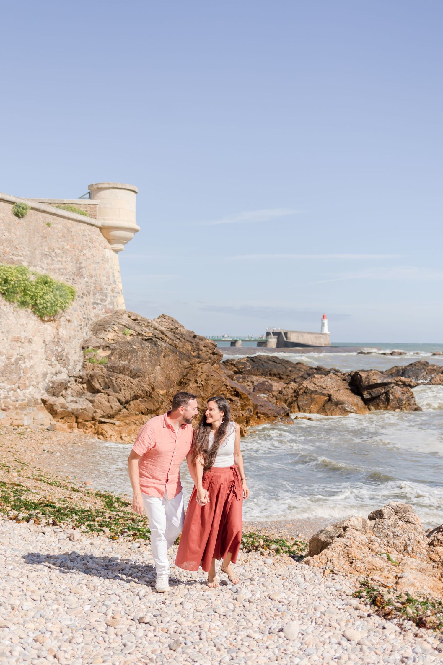 Two people walk along a rocky beach with a stone fortress wall and lighthouse in the background.