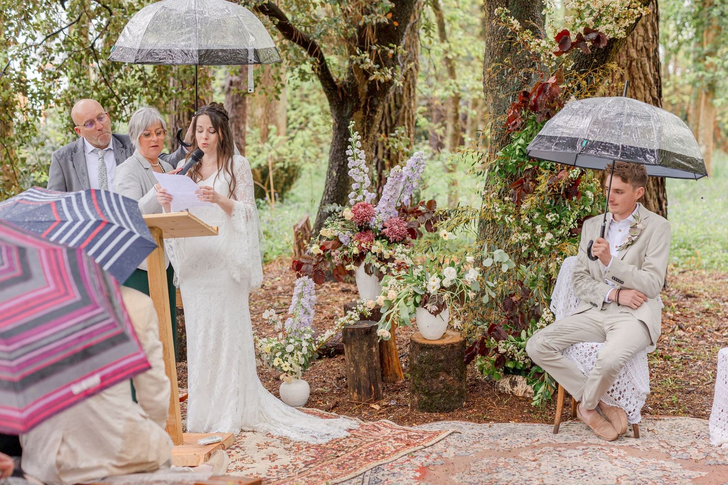 A wedding ceremony in a lush forest setting with clear umbrellas and floral decorations.