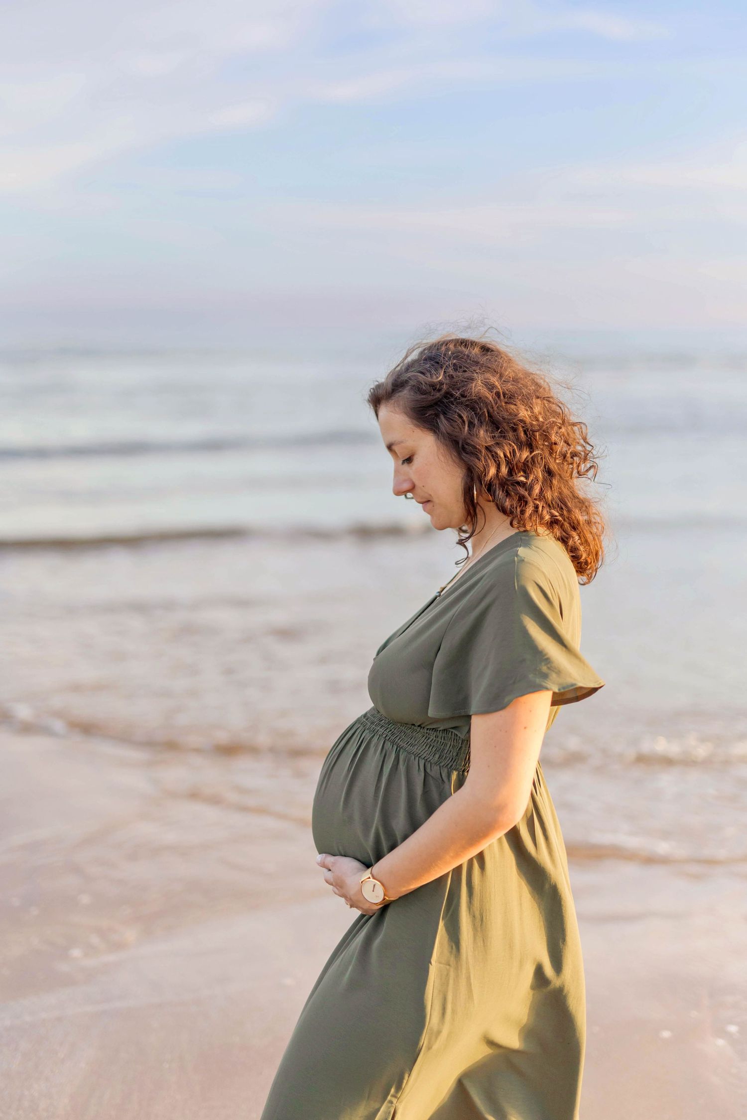 A person in a flowing green dress stands contemplatively on a beach at sunset.