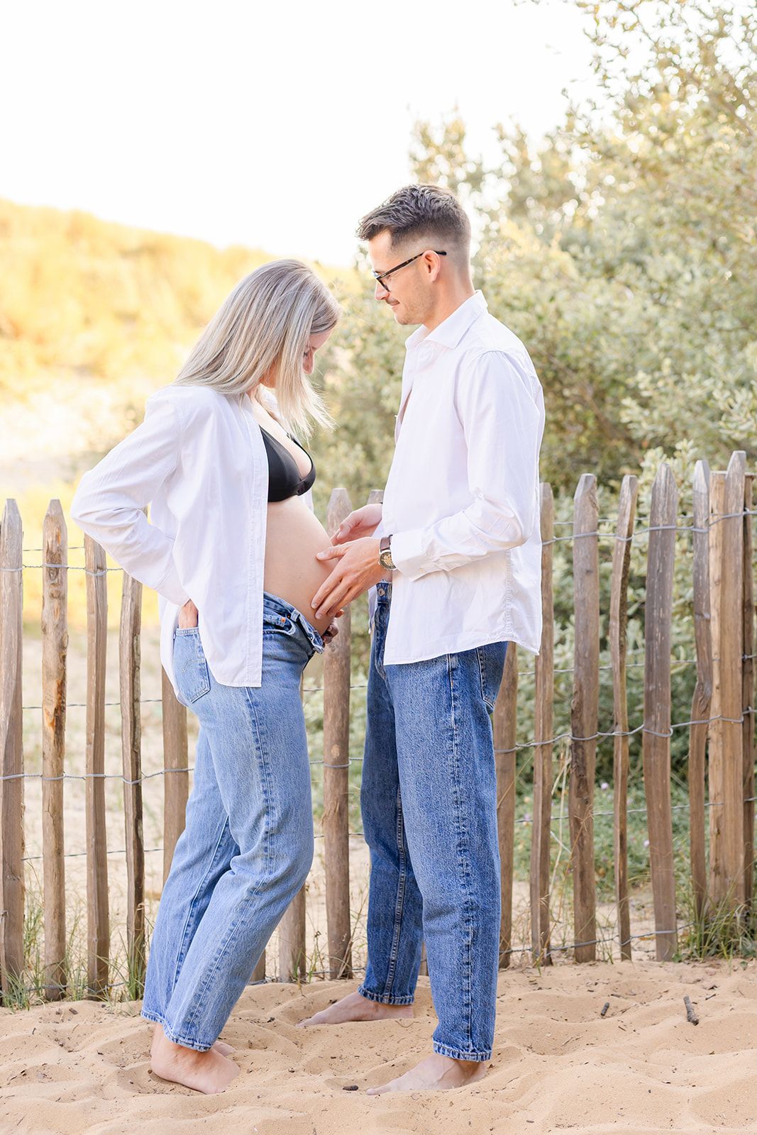 Couple in matching white shirts and jeans embrace on sandy beach near wooden fence at golden hour.