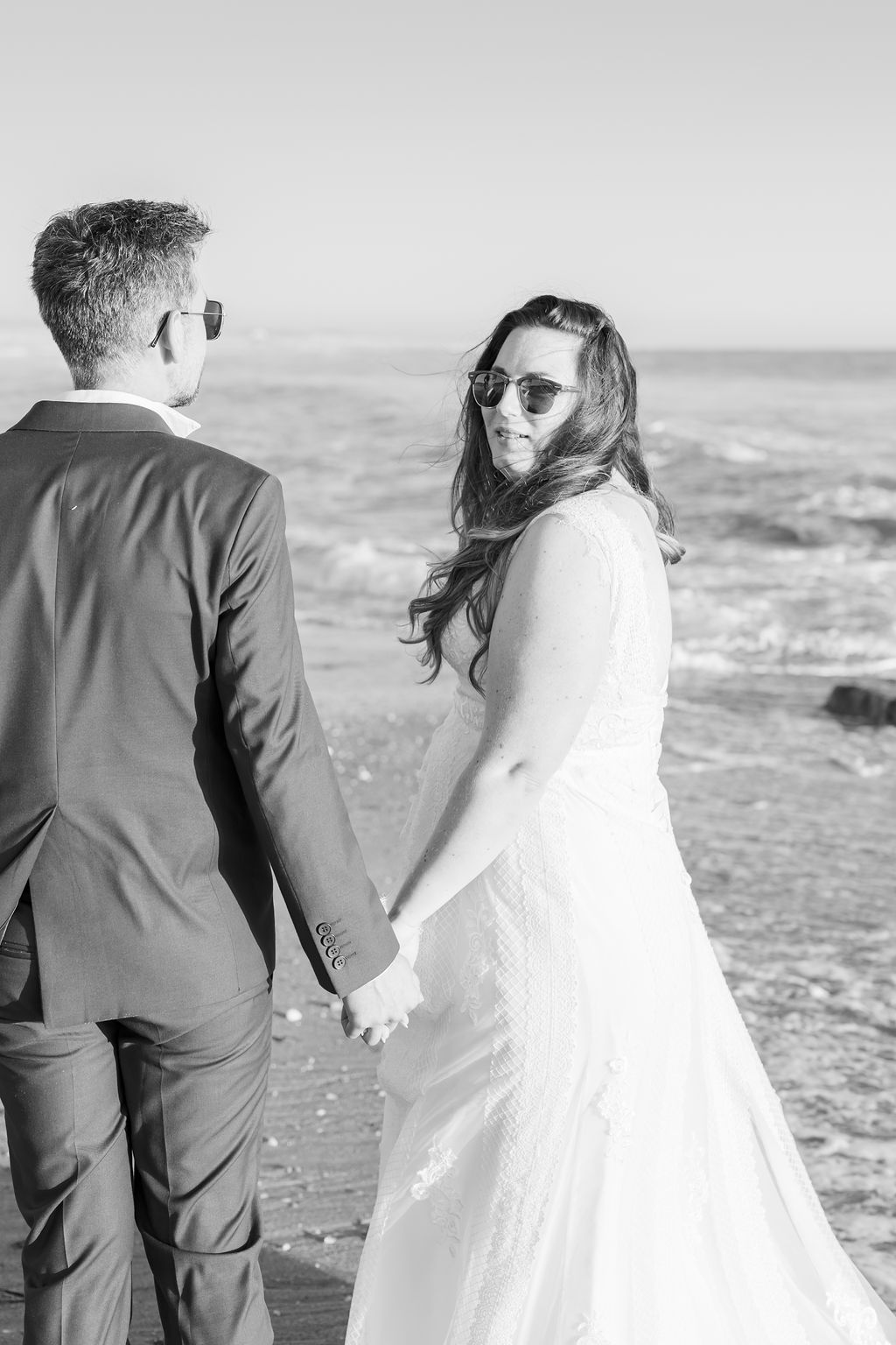 Black and white photo of couple holding hands while walking on beach.