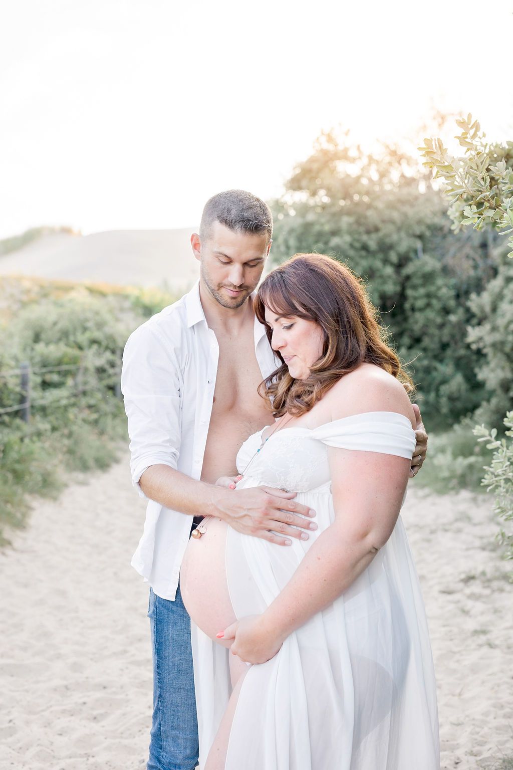 A romantic maternity photoshoot outdoors showing a couple embracing on a sandy trail surrounded by greenery at sunset.