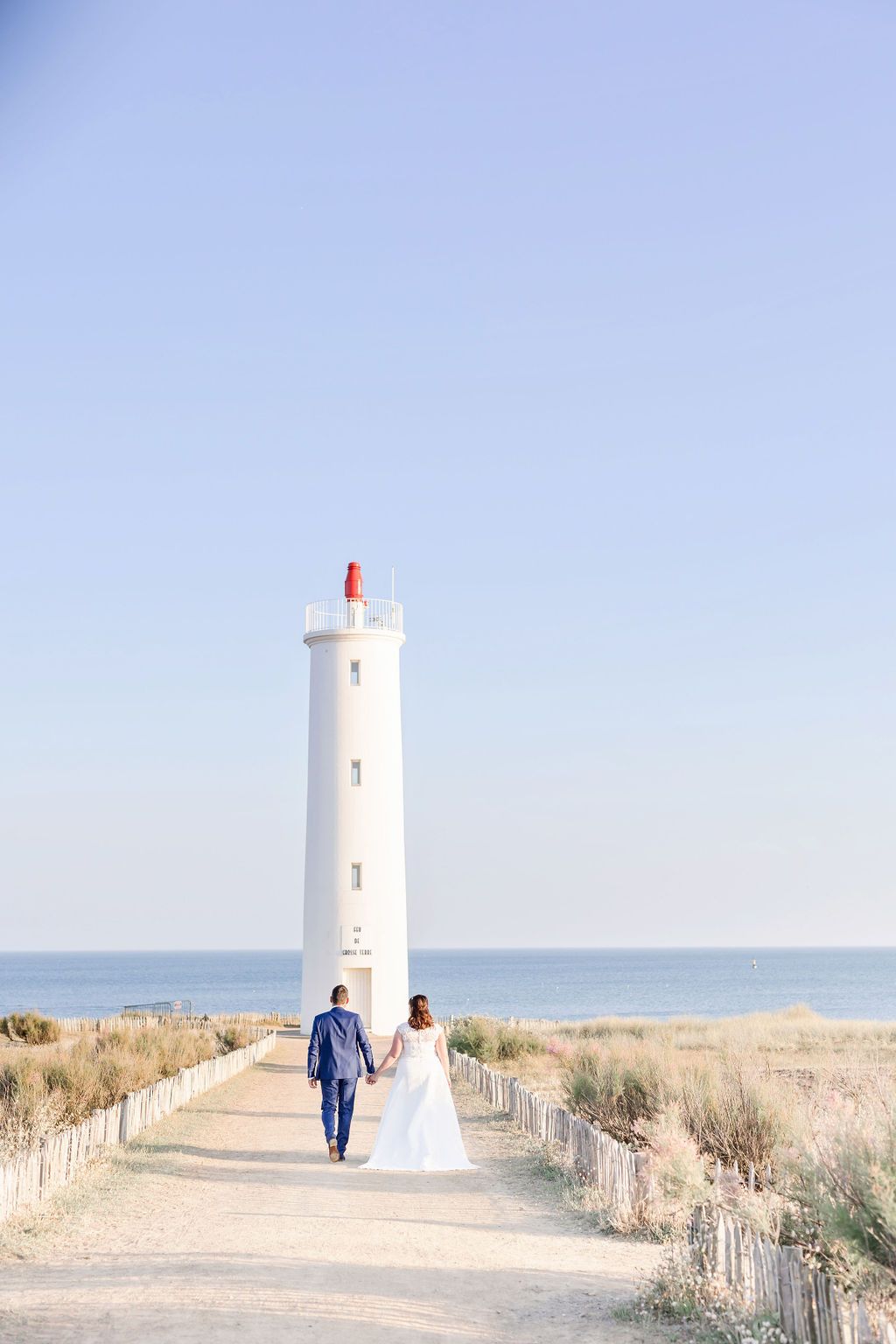 A couple walking toward a white lighthouse along a coastal path.