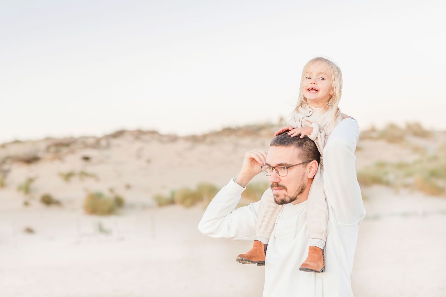 Mother and child in white dresses playing together on a serene sandy beach at sunset.