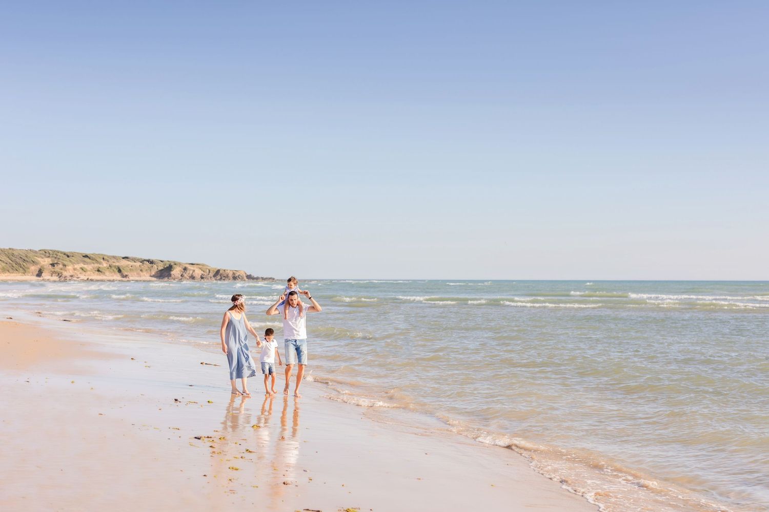 Family enjoying a sunny beach walk along the shoreline with gentle waves and coastal cliffs in the background.