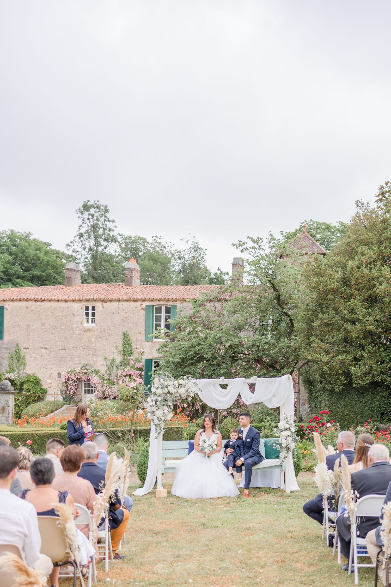 Outdoor wedding ceremony setup with white arch and chairs against historic stone building backdrop.