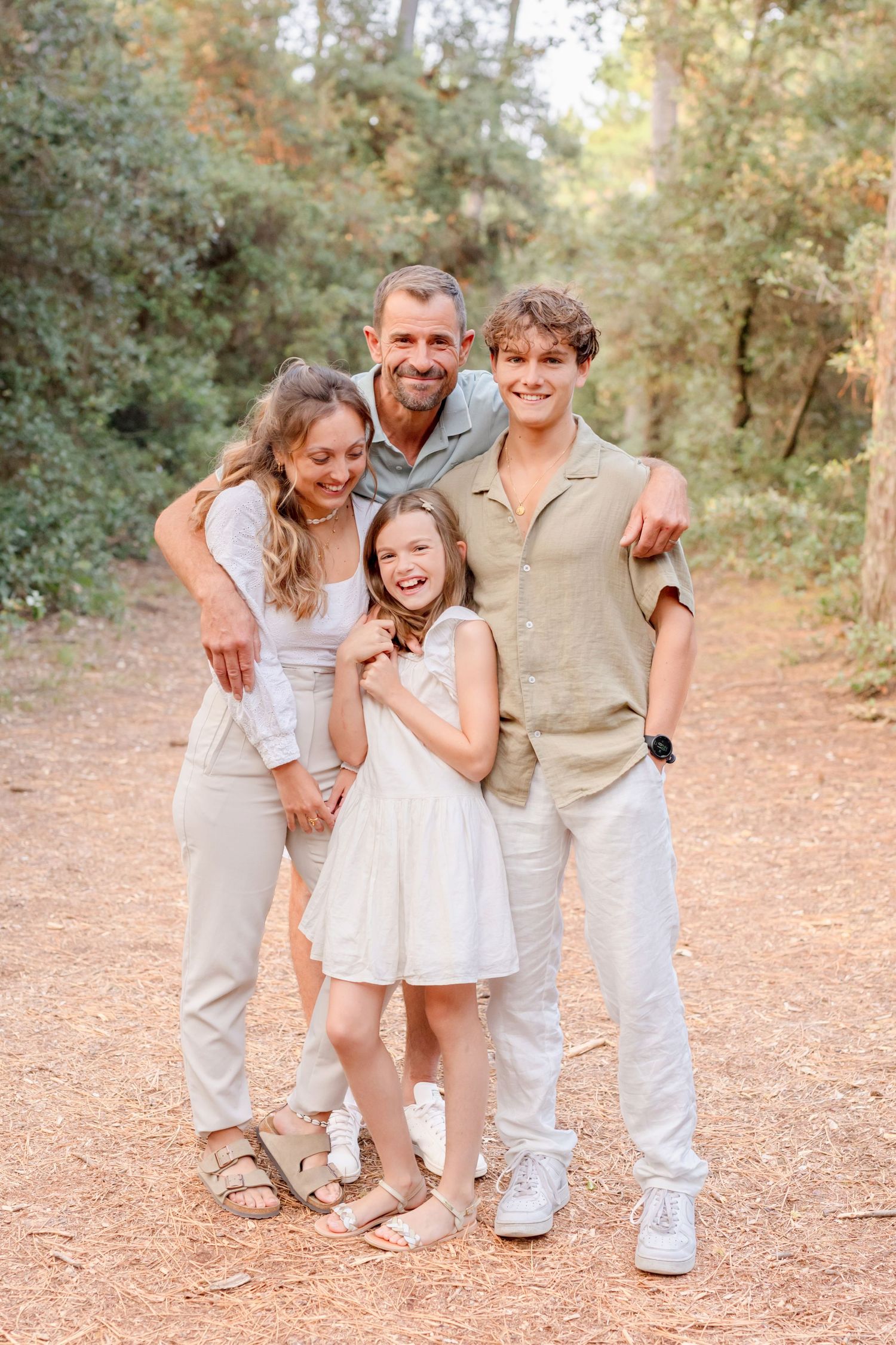 A happy family of four smiling together while standing on a forest path in neutral-toned clothing.