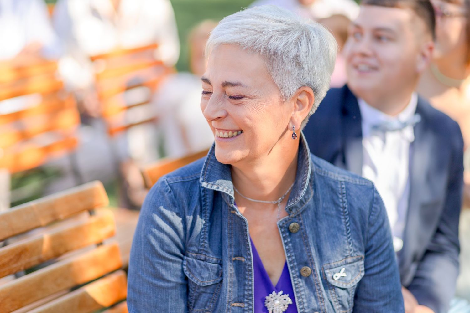 Wedding guest wearing denim jacket and purple top smiles joyfully while seated outdoors.