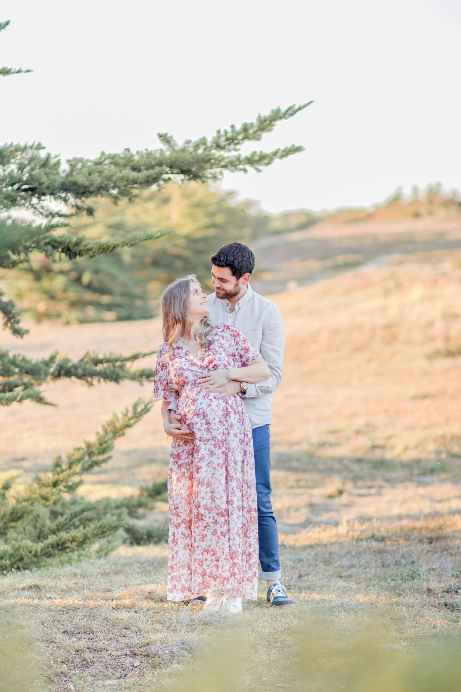 Couple embracing in a sunny field with pine trees in background.