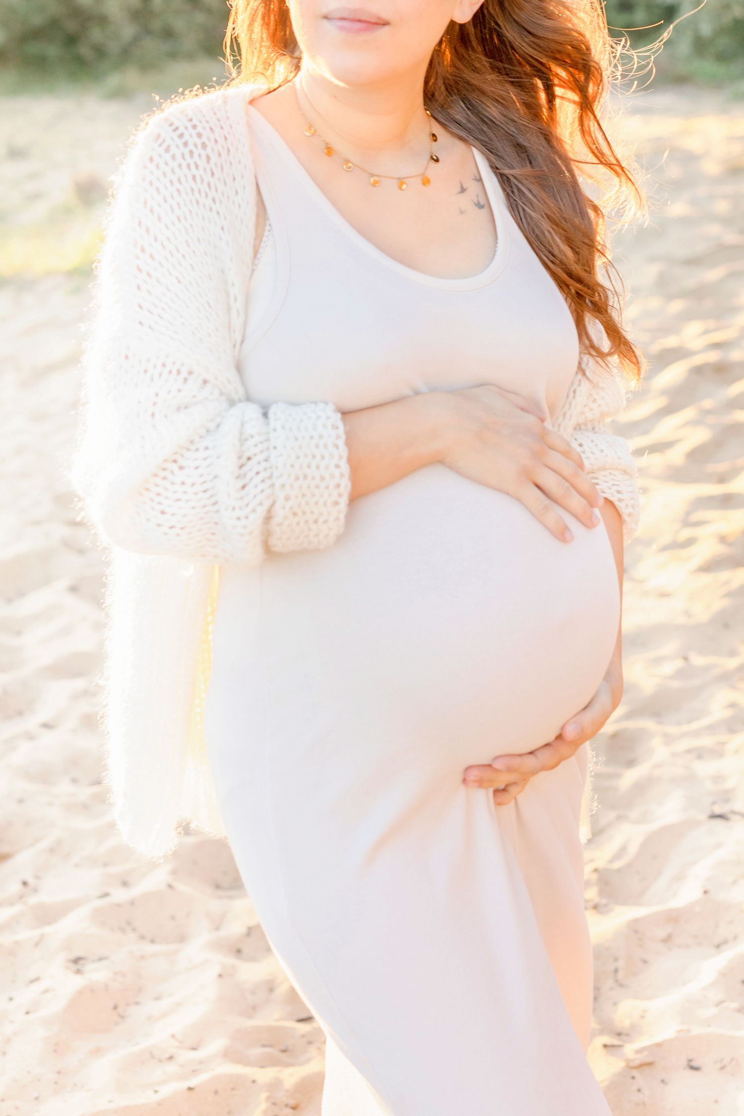 Expecting mother in white dress cradles baby bump during golden hour photoshoot on beach.