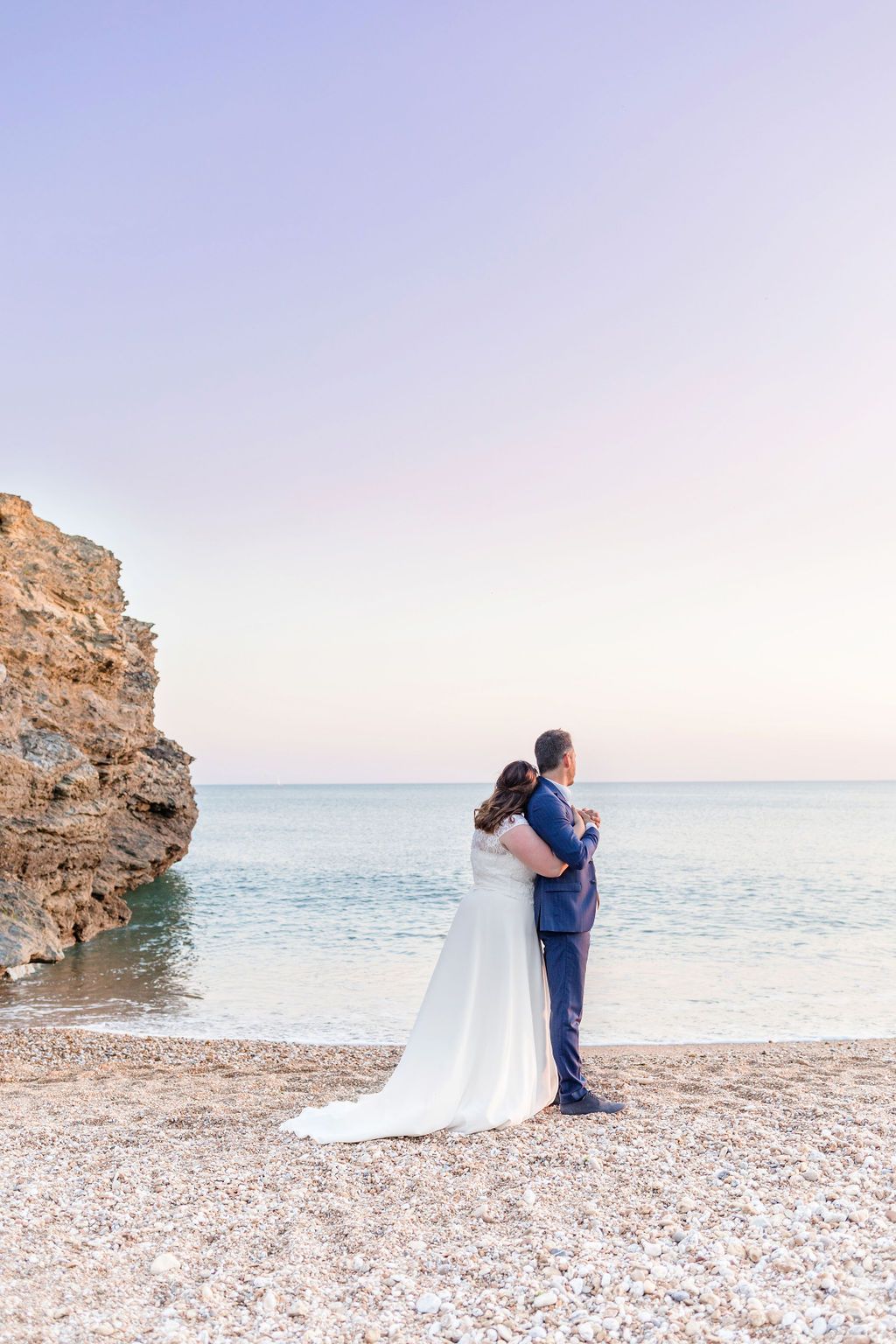 A wedding couple embraces on a scenic beach with rocky cliffs and crystal blue water.