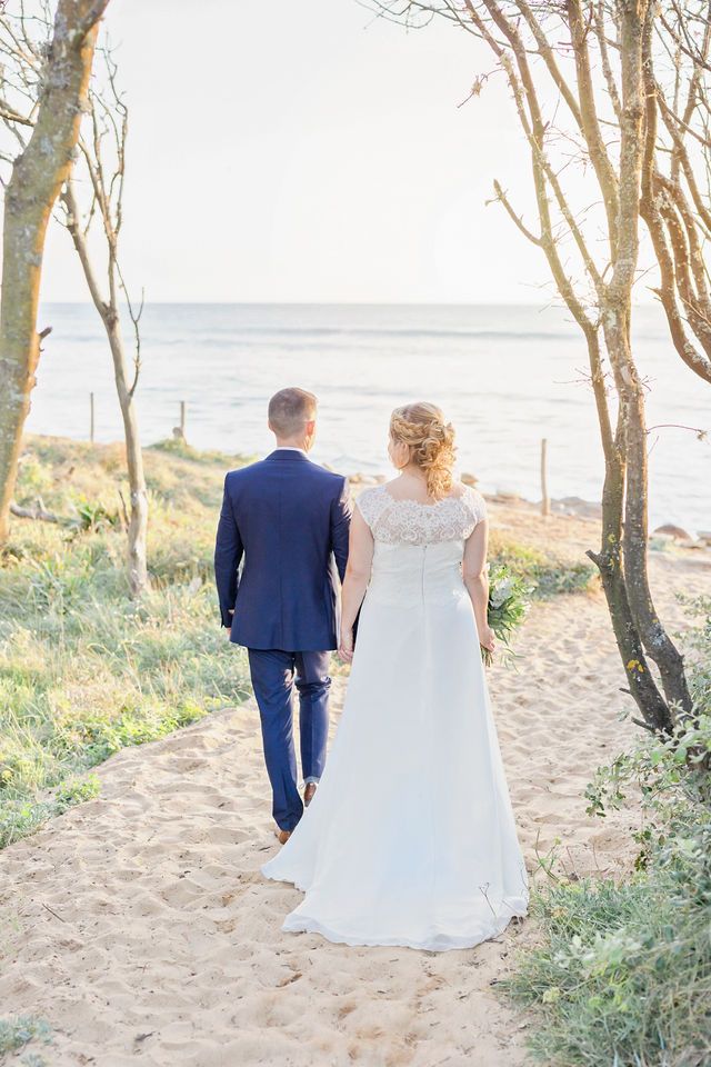 A couple in wedding attire walk along a sandy beach path lined with trees at sunset.