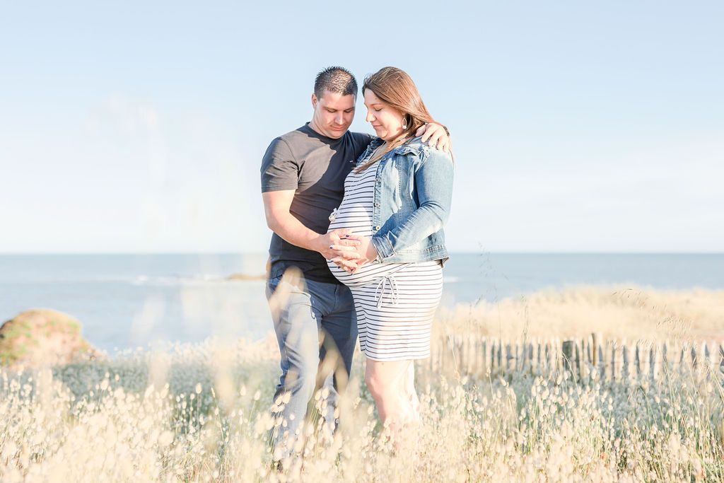 Couple embracing in seaside field with lighthouse in background.