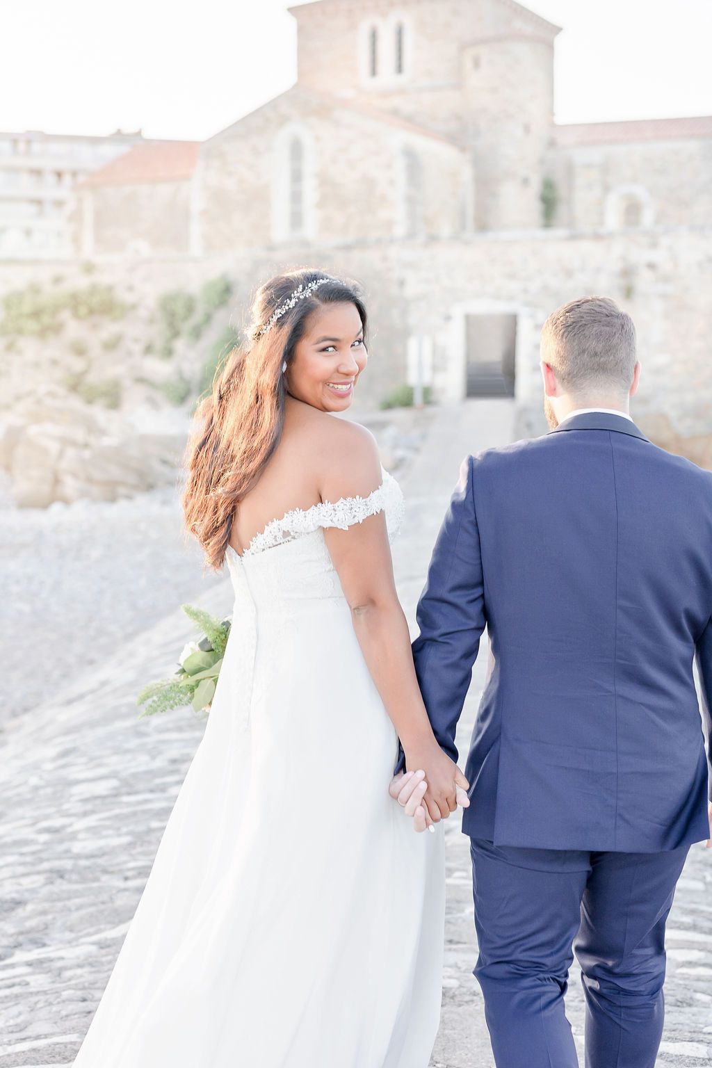 Bride in flowing white dress and groom in navy suit walking along cobblestone path.