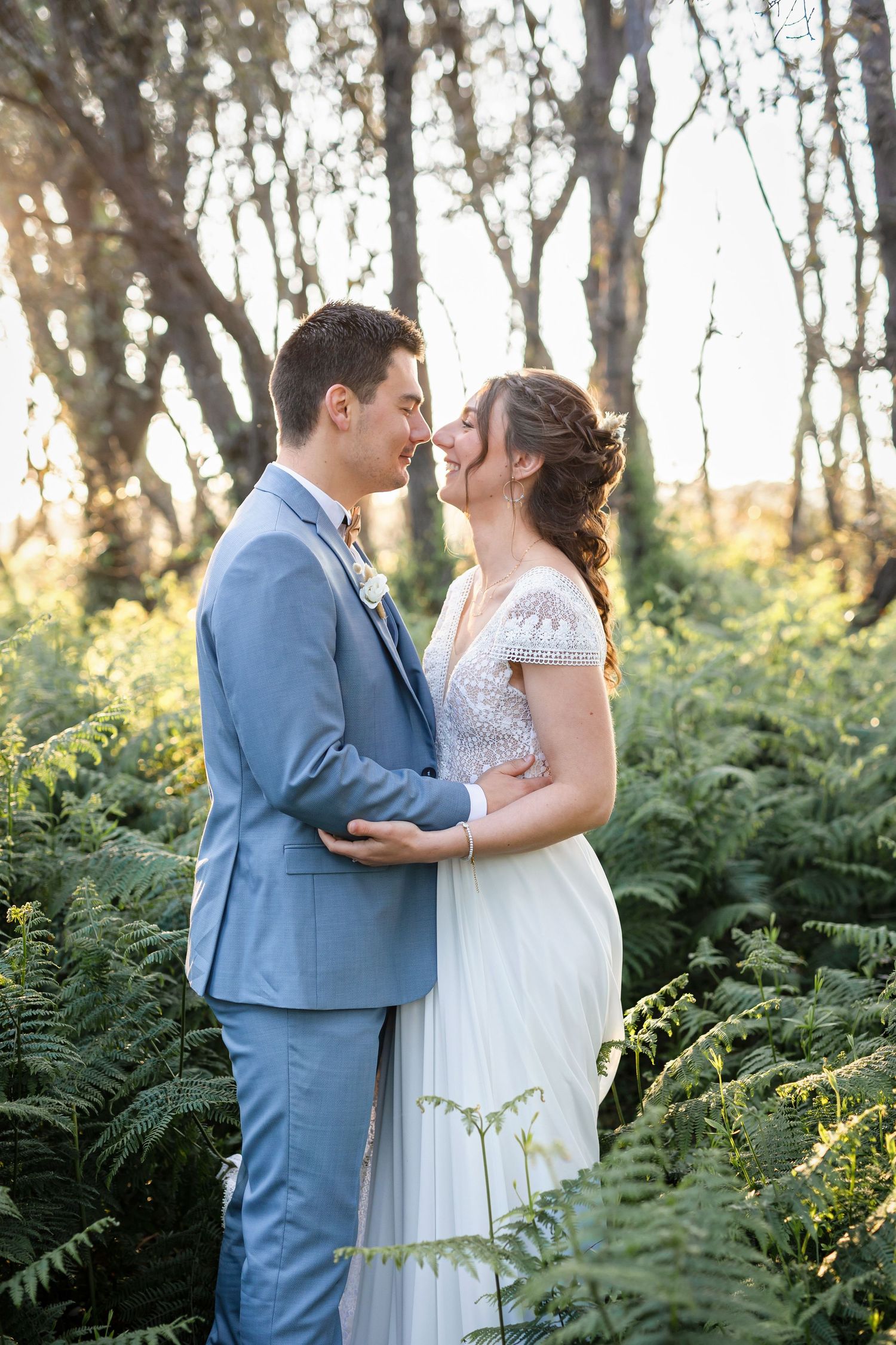A tender moment between newlyweds standing among ferns in a wooded area at sunset.