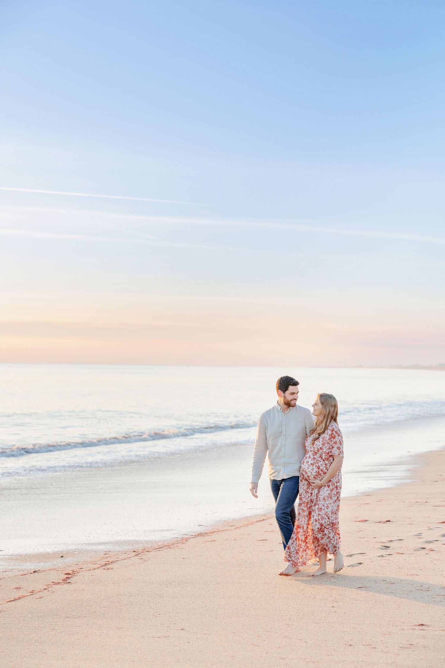 A couple strolls along a peaceful beach during sunset wearing white and floral clothing.